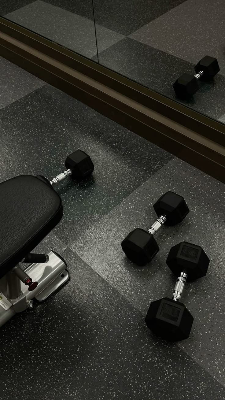Hex dumbbells and a bench on a speckled gym floor beside a mirrored wall.