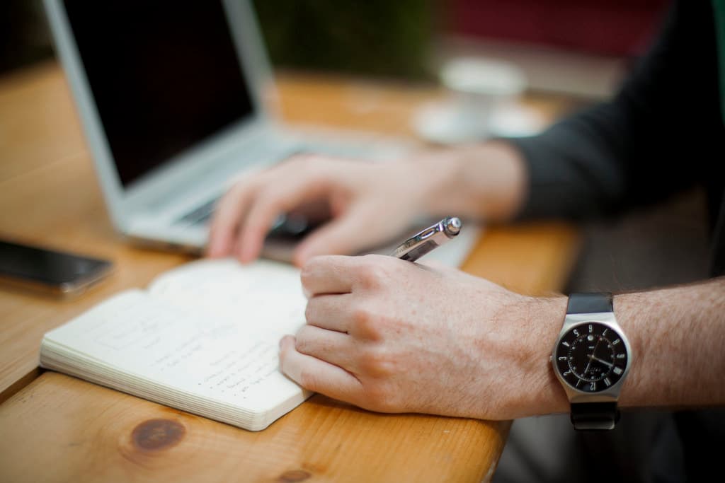 Writing in a notebook at a wooden desk, beside an open laptop.