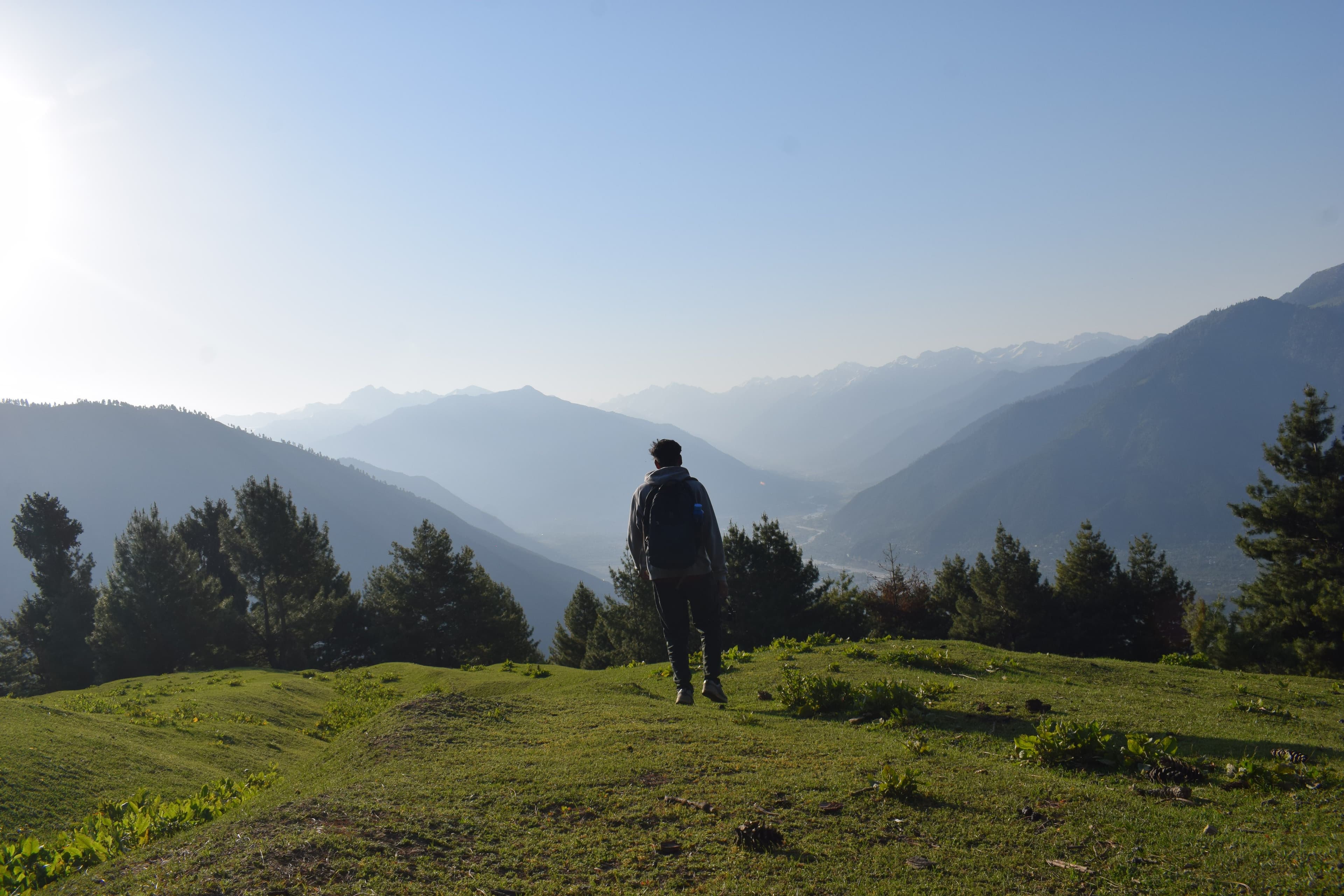 A young man in a white shirt standing at a hillside viewpoint, looking out over a green valley and the ridges behind it.