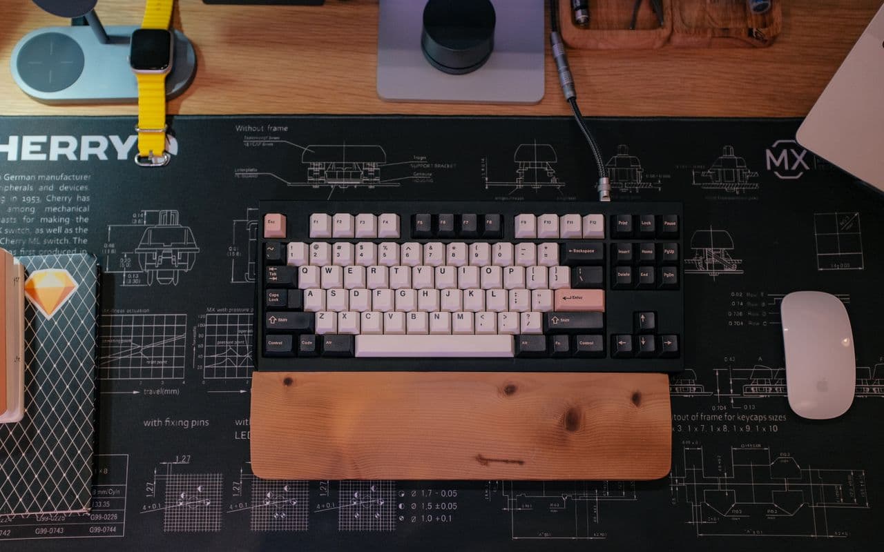A desk shot from above — a mechanical keyboard on a blueprint deskmat, wooden wrist rest, mouse and the edge of a laptop.